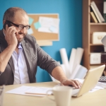 man on the phone with glasses smiling down with his hand on laptop keyboard